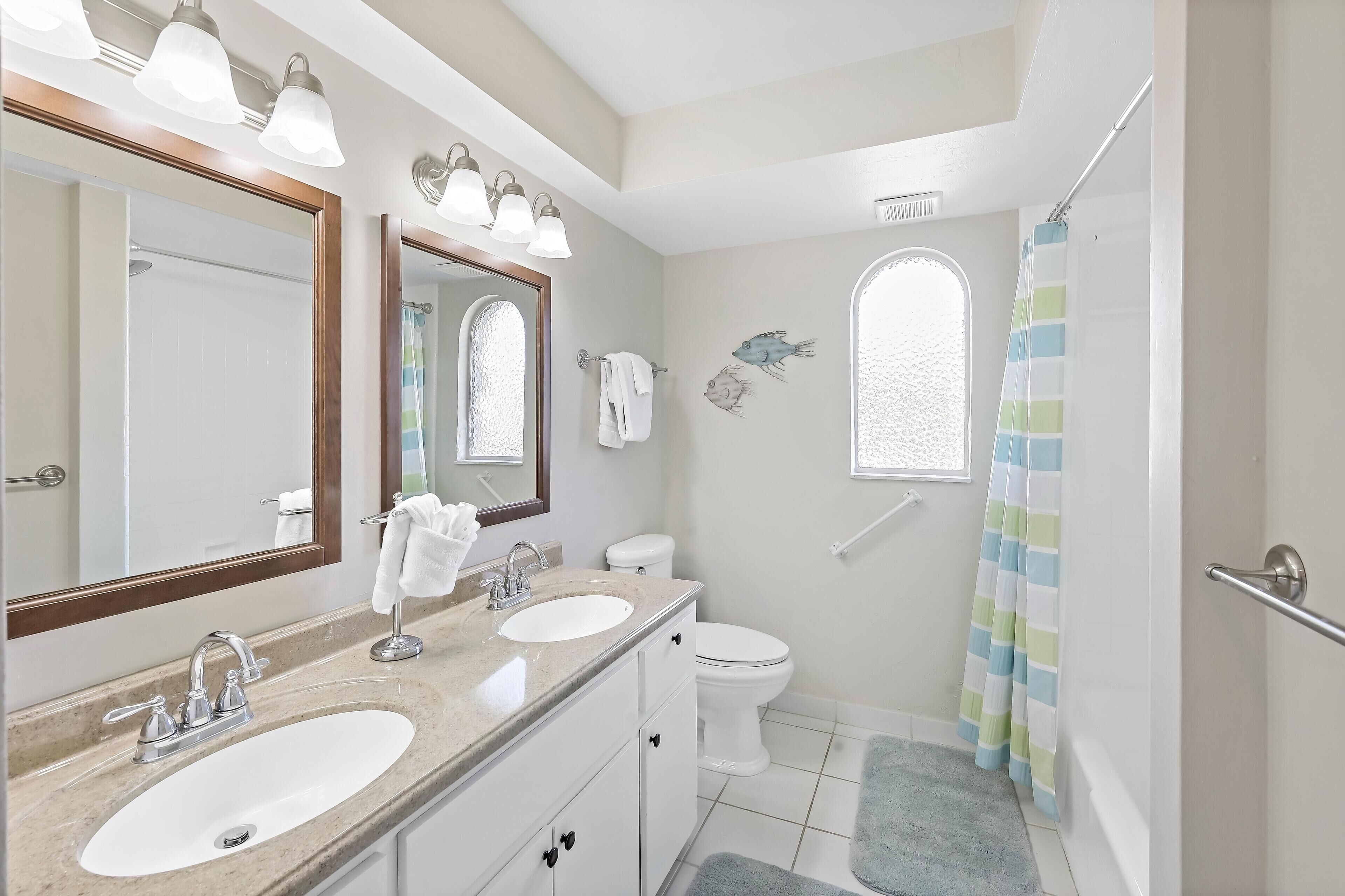 A bathroom with a double sink vanity, a toilet, a shower with a striped curtain, and a window with fish decals.