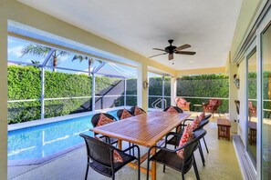 A sunlit patio with a pool, dining table, and chairs, overlooking a lush green landscape. - Pet-friendly Home With Pool and Steps to Beach/food (Marco Island)