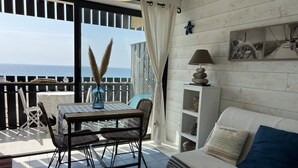 Una habitación con balcón, una mesa con sillas, un sofá, una lámpara y una estantería con objetos decorativos.