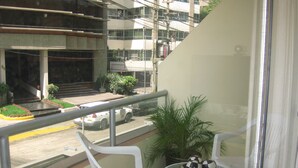 Uma varanda com duas cadeiras de plástico branco, uma mesa com objetos decorativos e vista para a rua, onde se vê um carro branco e um edifício ao fundo.