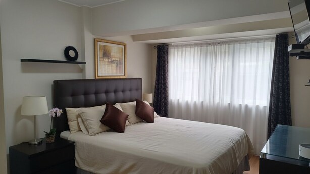 Quarto de hotel com uma cama grande, mesa de cabeceira com abajur, pequena planta em vaso, quadro na parede e janela com cortinas.