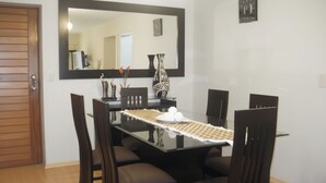 Uma sala de jantar com uma mesa retangular, seis cadeiras, um espelho sobre a mesa e um vaso com flores.