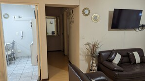 Uma sala de estar com um sofá de couro marrom, uma televisão de tela plana na parede e um corredor que dá para um banheiro.