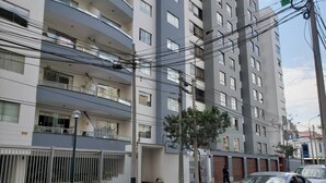 Um edifício residencial com várias andares, varandas, portão e uma rua com carros estacionados.