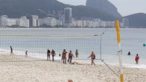 Un partido de voleibol de playa con una red en primer plano, una playa arenosa y un paisaje urbano con edificios y una montaña destacada al fondo.