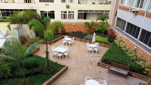 Un jardín en la azotea con varias mesas y sillas blancas, rodeado de abundante vegetación y plantas en macetas.