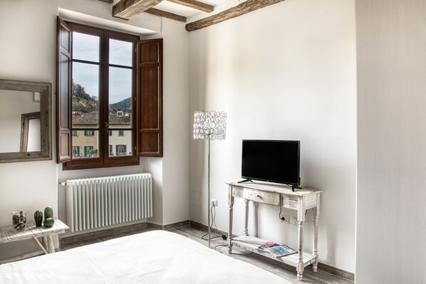 A bedroom with a bed, a dresser, a television on a stand, a radiator, and a window with a view of buildings. - Il Nido in Chianti (Greve in Chianti)