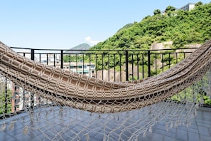 A hammock on a balcony with a view of a green hillside and buildings in the background.