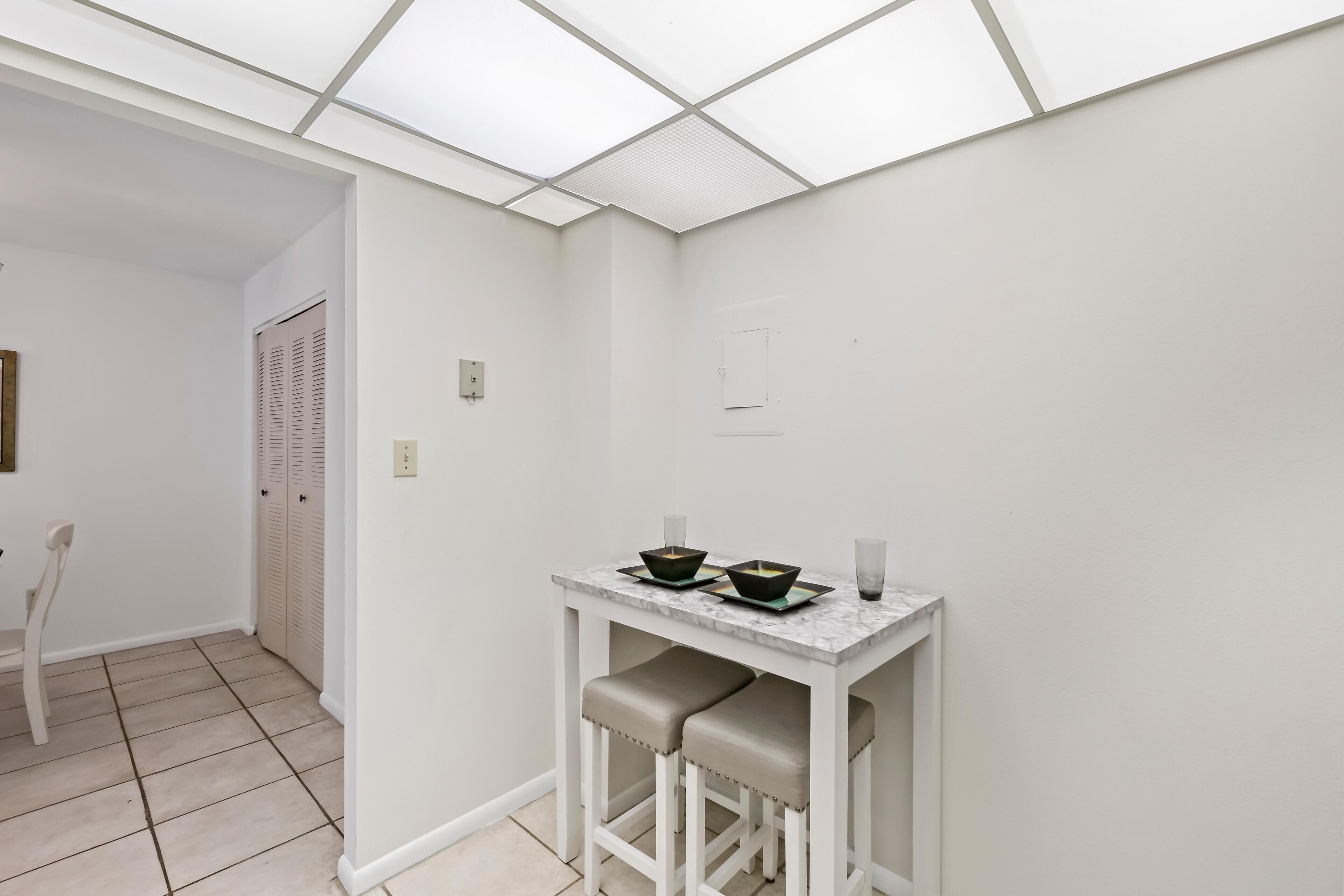 A small, white room with a tiled floor, a white table with two stools, and a door with louvered shutters.