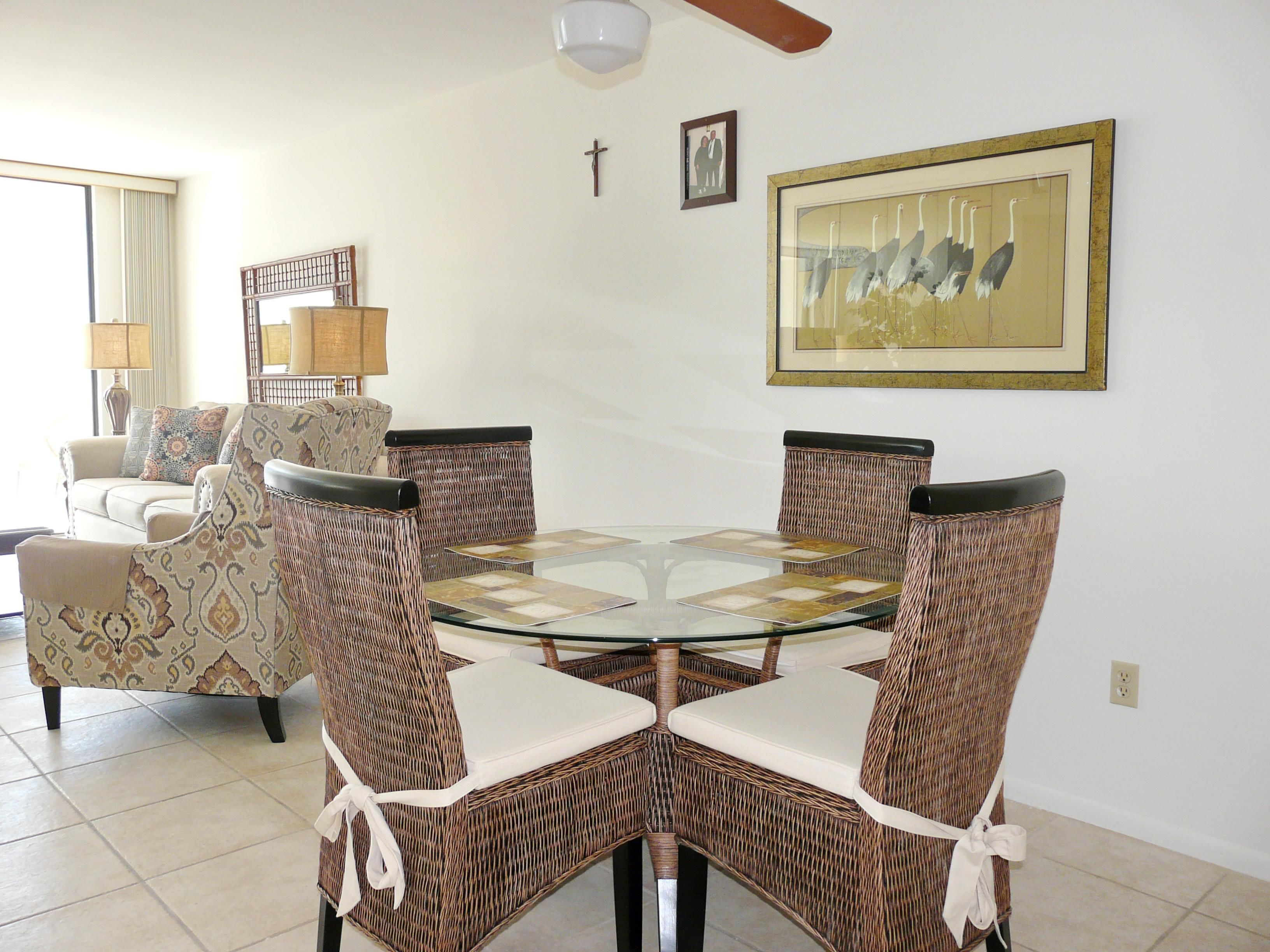A dining area with a glass-top table surrounded by wicker chairs with white cushions. There is a sofa and a lamp in the background.