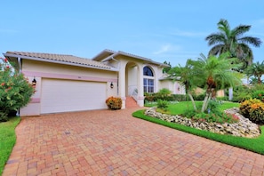 A single-story house with a garage, a driveway, and a well-maintained garden with palm trees and flowering plants. - 910 Olive Court (Marco Island)