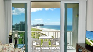 A balcony with a view of the beach and ocean, white wicker furniture, and a pool. - Sunny Beachfront Condo With Pool and Sw-facing Balcony (Marco Island)