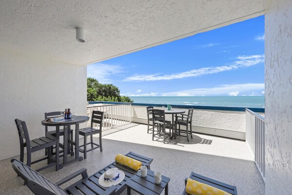 A balcony with a view of the ocean, furnished with tables and chairs, and a small table with a bottle and glasses. - Unmatched Beachfront Condo With Roomy Balcony and Pool (Marco Island)