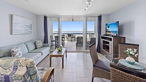 A modern living room with a sofa, coffee table, and wicker chairs. A television is on a wooden cabinet, and there is a view of the beach through large windows. - Unmatched Beachfront Condo With Roomy Balcony and Pool (Marco Island)