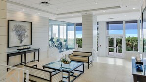 A spacious, modern lobby with a glass wall offering a view of the outdoors, a seating area with a glass coffee table, and a framed artwork on the wall. - Beachfront Condo With 180-degree Views and Pool (Marco Island)