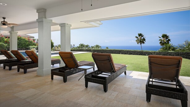 Ein überdachter Außenbereich mit Korbmöbeln und Deckenventilator, mit Blick auf einen angelegten Garten und das Meer.
