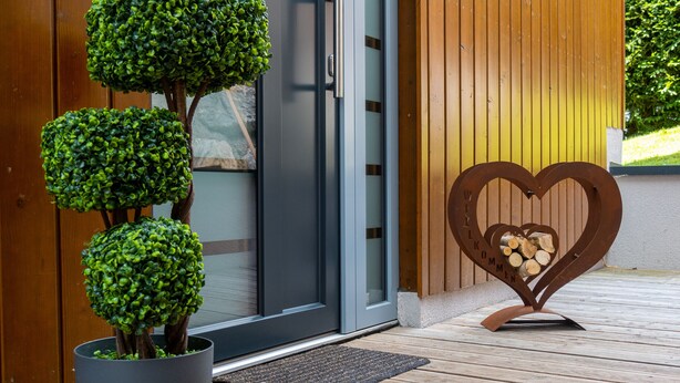 A modern entrance with a potted plant, a heart-shaped welcome sign, and a wooden deck. - Schmitten Finest Apartment - SKYBLUE (Zell am See)