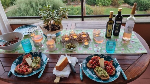 A dining table with plates of food, glasses, and bottles of wine, set on a balcony with a view of greenery.
