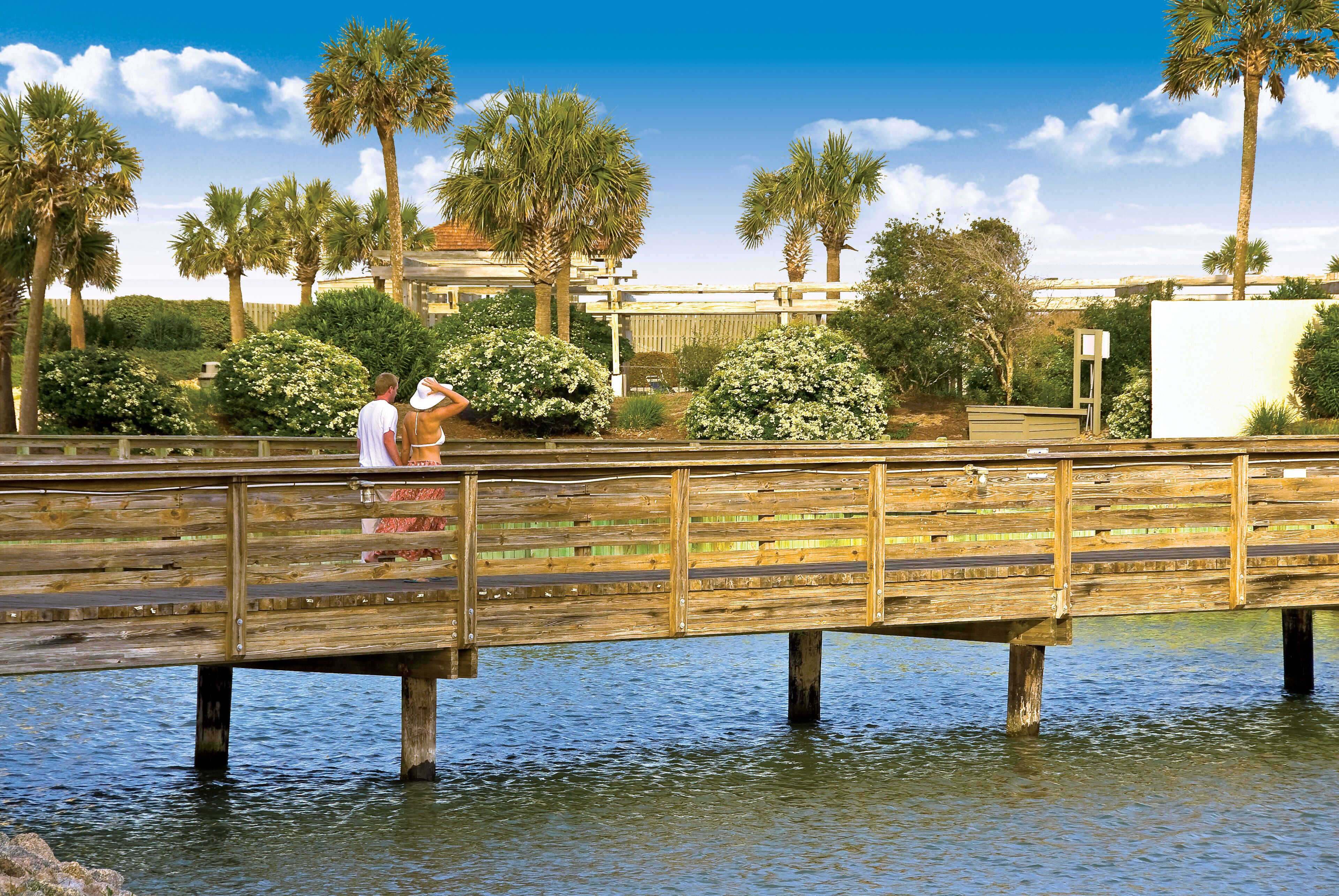 A wooden bridge over water with palm trees and a building in the background.