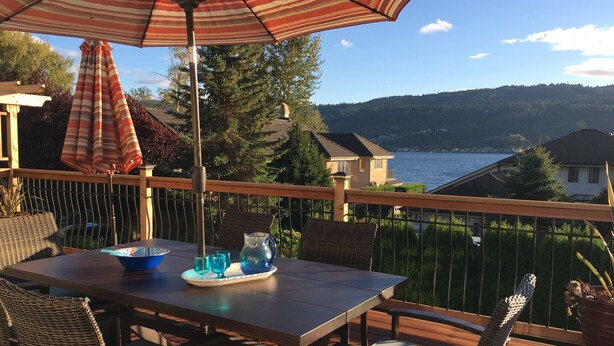 Una terraza de madera con una mesa y sillas, un parasol a rayas y una vista panorámica de un lago y colinas.