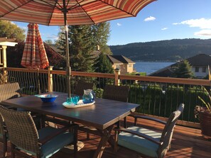 Una terraza de madera con una mesa y sillas, un parasol a rayas y una vista panorámica de un lago y colinas.