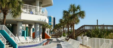 A resort with a pool, lounge chairs, palm trees, and a staircase leading to a balcony.
