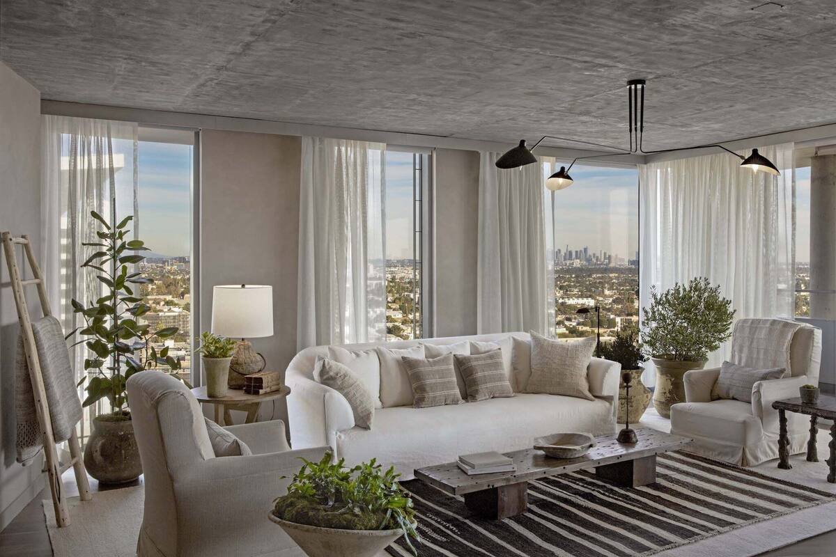 Un salon moderne avec un canapé blanc, des fauteuils et une table basse. De grandes fenêtres sont ornées de rideaux légers, et l’on peut admirer une vue sur le paysage urbain, ainsi qu’une plante en pot.