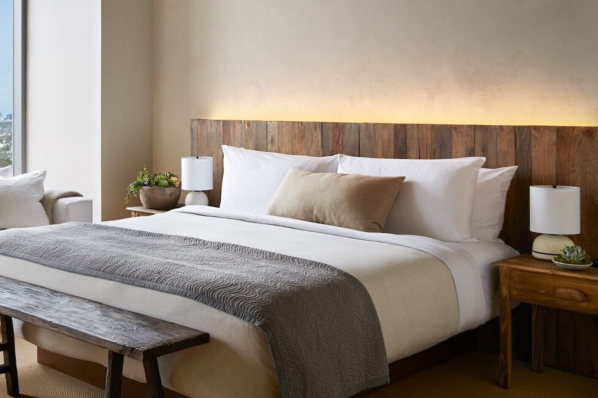 Un lit bien fait, avec une tête de lit en bois, une literie blanche et un plaid gris. Il y a deux tables de chevet, chacune surmontée d’une lampe et d’une petite plante. Un banc en bois est placé devant le lit.