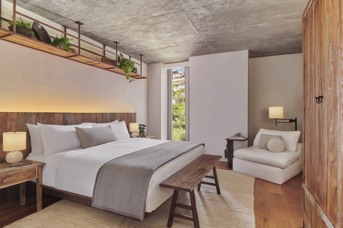 Une chambre avec un grand lit, des tables de chevet, un banc, un canapé et une vue sur la verdure par la fenêtre.