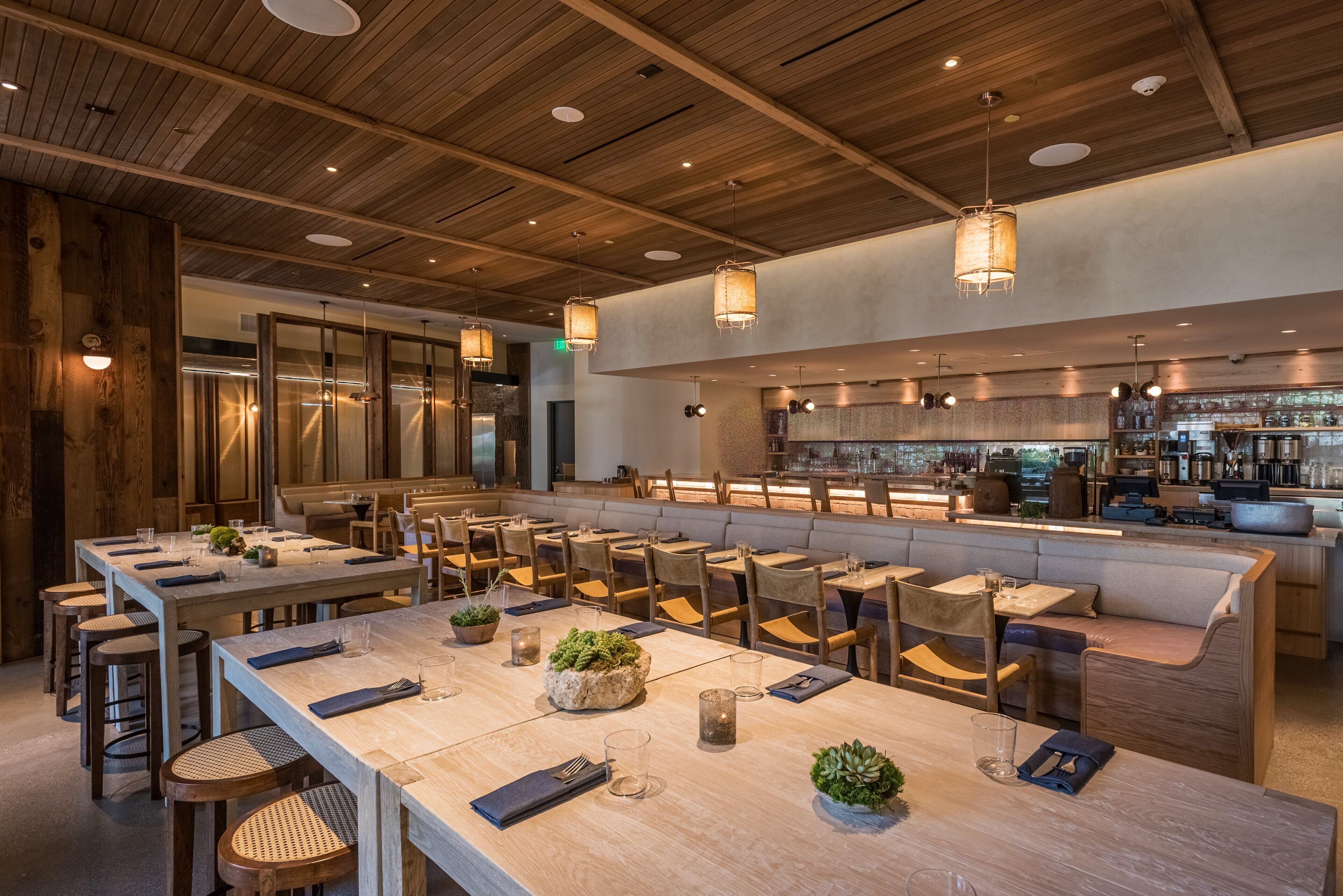 A modern restaurant with wooden ceiling beams, long communal tables, and a bar area.
