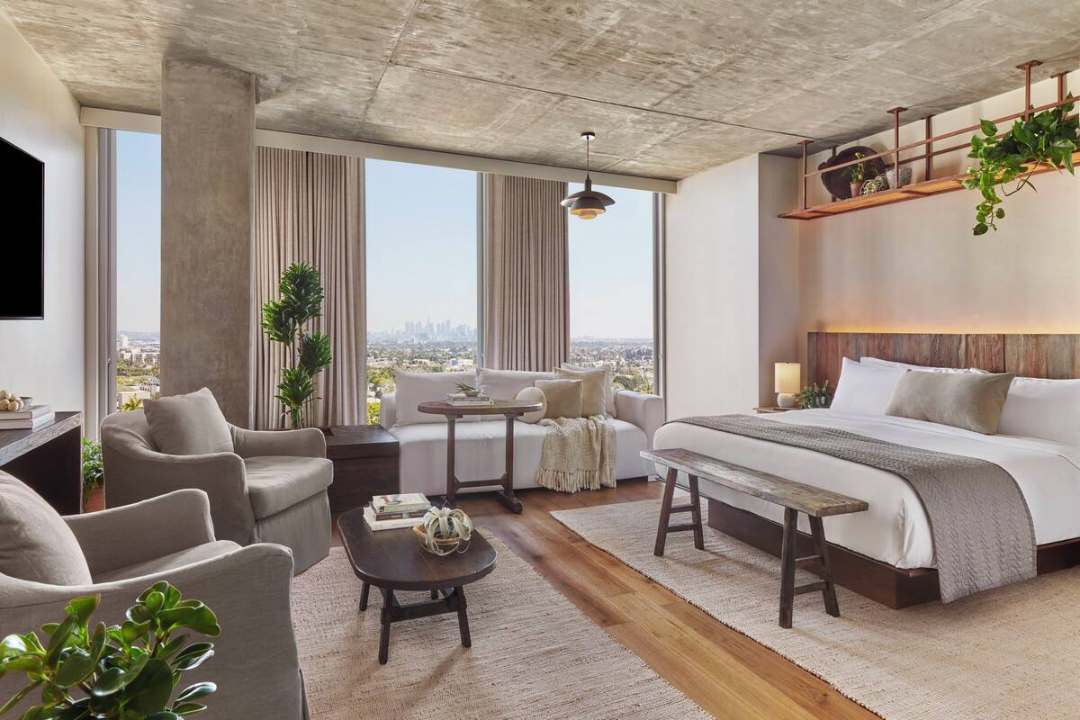Une chambre d’hôtel moderne, dotée d’un grand lit, d’un canapé, d’une table basse et d’un bureau. De grandes fenêtres offrent une vue sur la ville.