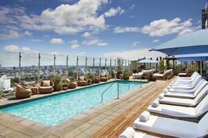 Piscina en la azotea con sillas de descanso y plantas en macetas, con vista a un paisaje urbano.
