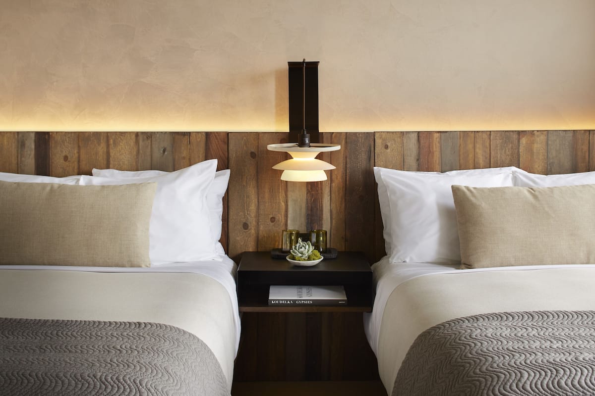 Une chambre d’hôtel avec deux lits, une tête de lit en bois, une table de chevet avec une lampe et un livre intitulé « KOUDELKA GYPSIES ».