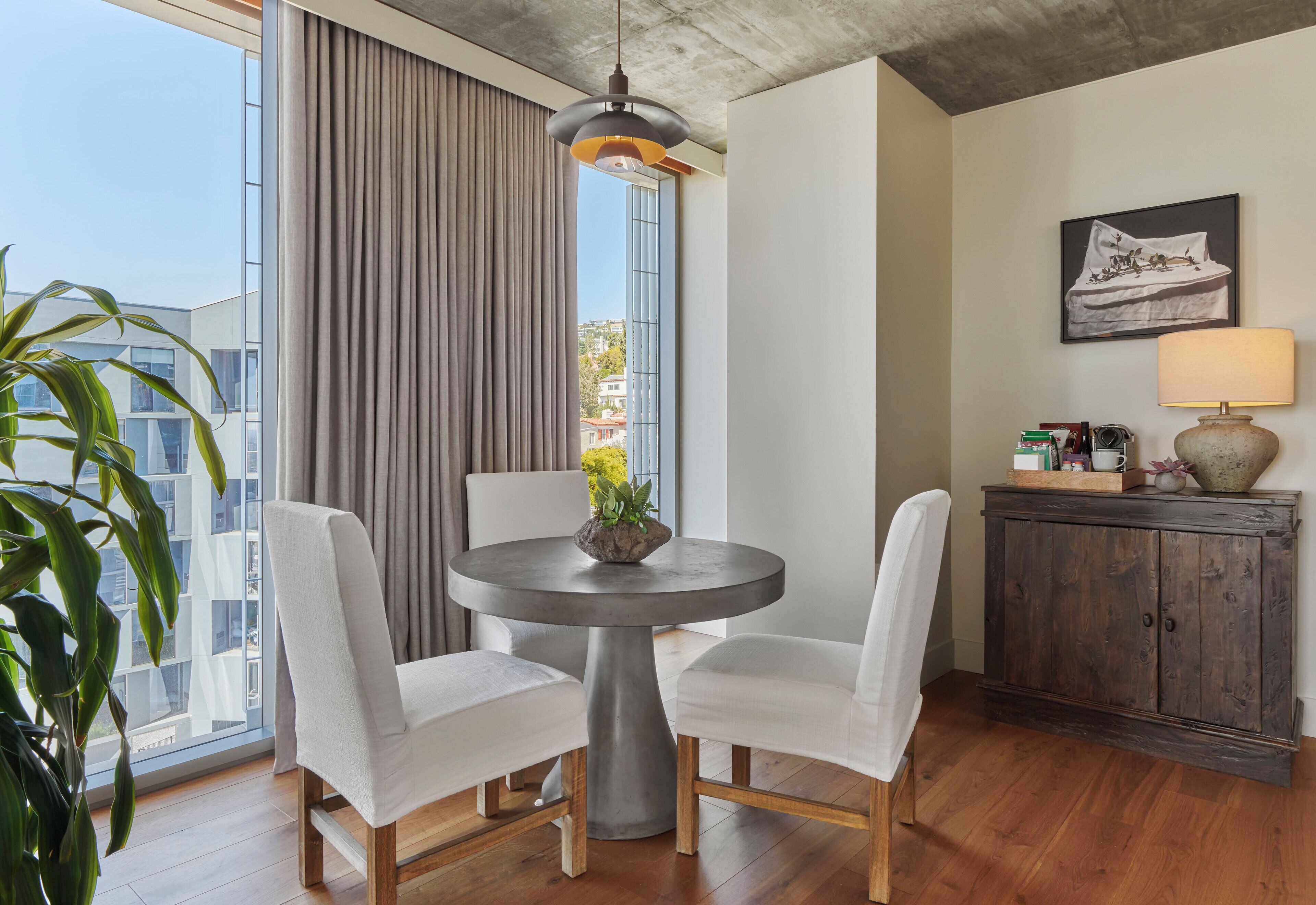 Ein modernes Zimmer mit einem runden Esstisch, zwei weißen Stühlen, einem Blumentopf, einem Holzschrank und einem großen Fenster mit Vorhängen.