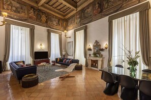 A spacious living room with a large sofa, a coffee table, and a TV. There is a fireplace with a mantel and a dining table with chairs. The room has ornate ceiling artwork and large windows with curtains.
