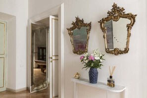 Eine offene Tür führt in einen Raum mit Marmorboden, einem weißen Konsolentisch mit Blumenvase und zwei verzierten Spiegeln an der Wand.