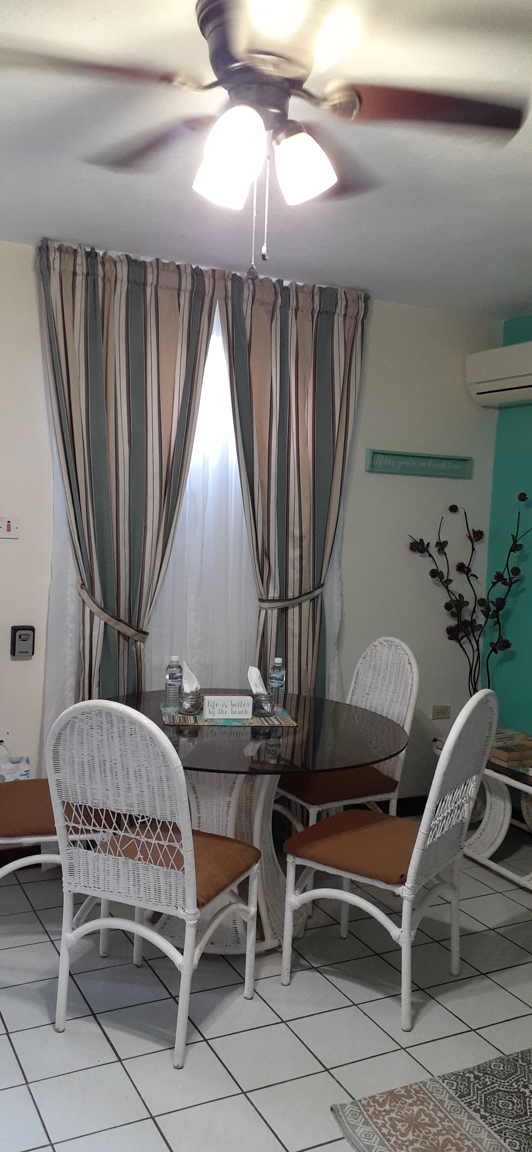 A dining area with a round glass table, white wicker chairs with brown cushions, striped curtains, and a ceiling fan.
