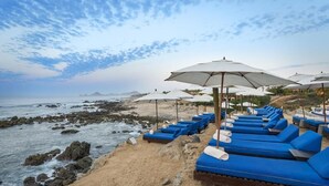 A beachside lounge area with blue lounge chairs and white umbrellas, overlooking a rocky shoreline and the ocean.