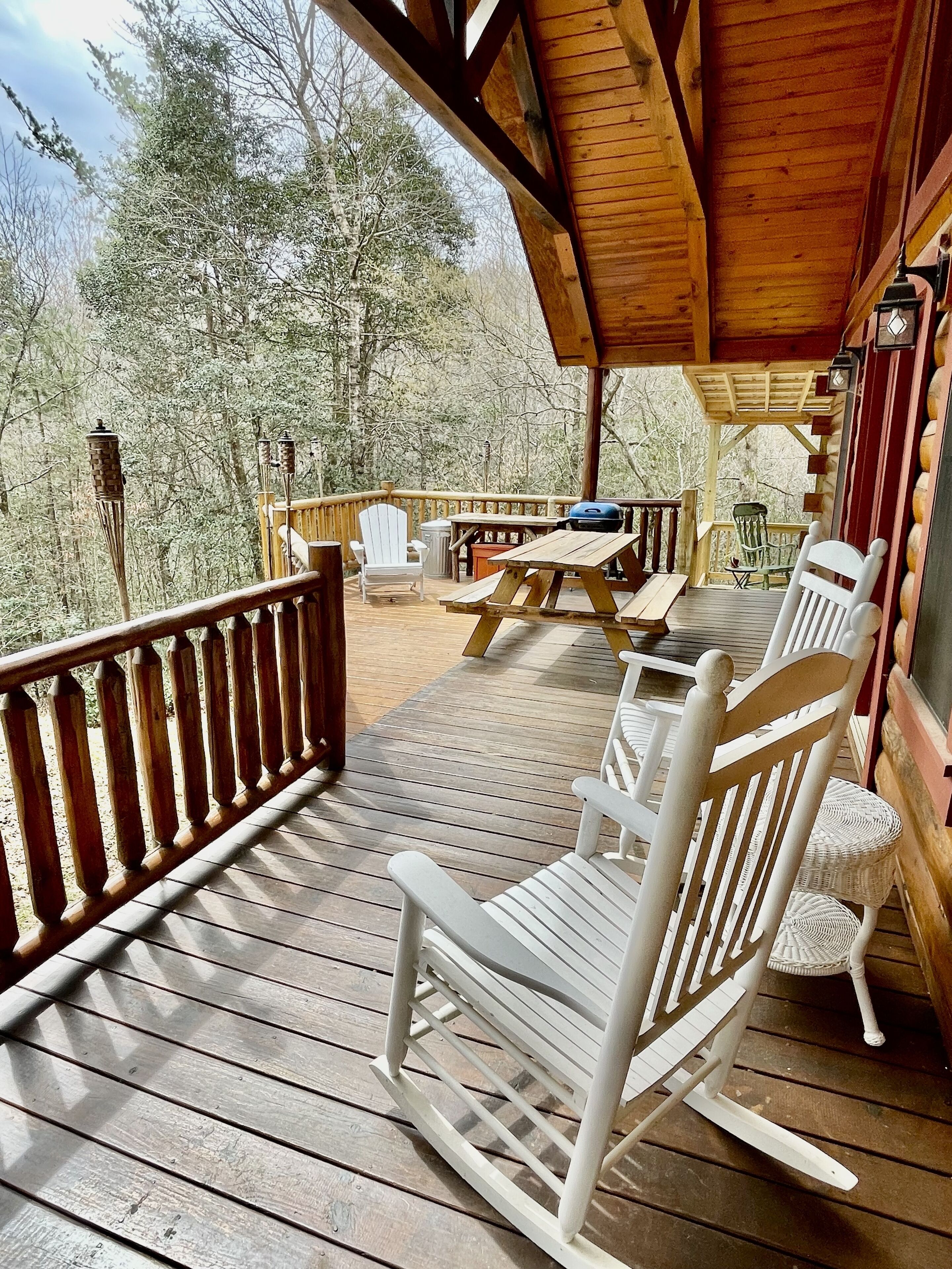 A wooden deck with white rocking chairs and picnic tables, surrounded by trees.