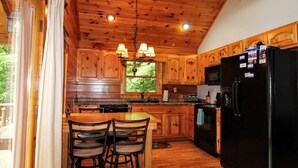A wooden cabin kitchen with a dining area, wooden cabinets, a black refrigerator, and a window with a view of greenery. - DAYBREAK LOG GETAWAY - NEW HOT TUB! (Murphy)