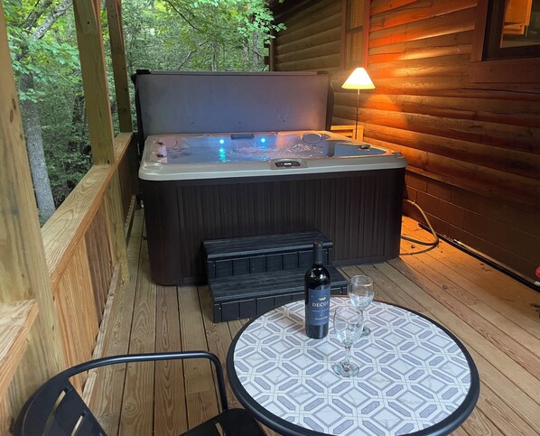 A hot tub with a wooden deck, a table with a patterned tablecloth, and a bottle of wine. - DAYBREAK LOG GETAWAY - NEW HOT TUB! (Murphy)