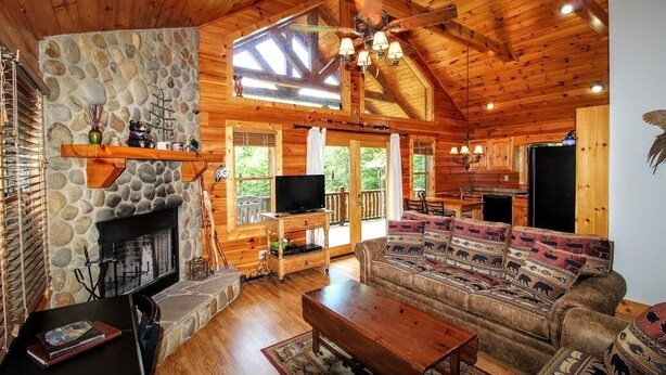 A cozy cabin interior with a stone fireplace, wooden ceiling, and a large sofa. - DAYBREAK LOG GETAWAY - NEW HOT TUB! (Murphy)