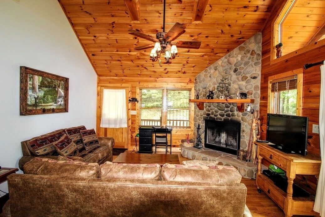 A cozy cabin interior with a stone fireplace, wooden ceiling, and a television on a wooden stand.