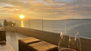 En balkong med ett glasbord och två sittplatser med bekväma dynor, med utsikt över en solnedgång över havet.