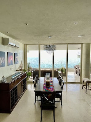 Enjoy meals with a view from this dining area, featuring a large table for four and a sliding glass door leading to a balcony overlooking the sea.