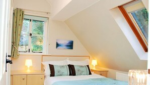 Een slaapkamer met een schuine kap, een bed met een blauwe sprei, twee bedlampjes, een raam met gordijnen en een schilderij aan de muur.