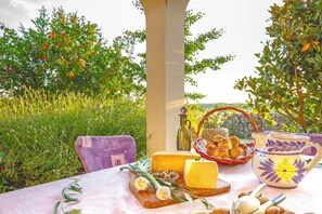 Ein Tisch im Freien mit Käse, Oliven, Brot und einer Krug, umgeben von Grünpflanzen und Bäumen.