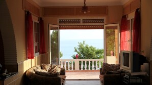 Een woonkamer met een bank en een televisie, een balkon met uitzicht op zee, en een rood gordijn aan het raam.