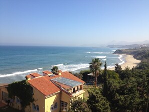 Une vue côtière avec une plage, des vagues de l’océan et un bâtiment doté d’un toit recouvert de tuiles rouges et de panneaux solaires.