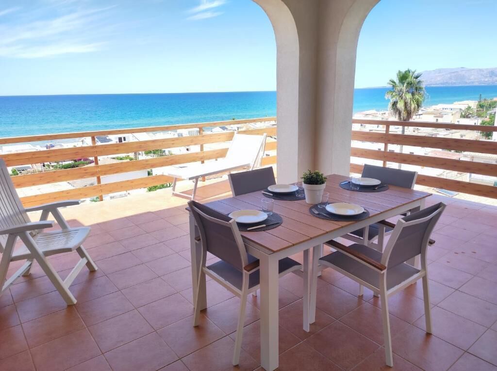 A terrace with a table set for two, overlooking a beach and mountains.